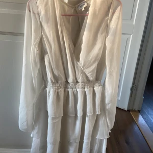 Vit omlottklänning med volanger från bubbleroom - Supersöt vit omlottklänning från bubbleroom med långa ärmar och volangdetaljer. Klänningen har resår i midjan och v-ringning framtill. Perfekt för sommarens alla tillfällen!