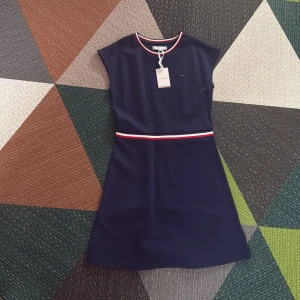 Marinblå klänning från Tommy Hilfiger - Snygg marinblå klänning från Tommy Hilfiger med sportiga ränder i rött, vitt och blått vid halsringning och midja. Klassisk och stilren design med rund hals och ärmlös modell. Perfekt för dig som gillar enkel men trendig stil.