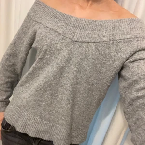 Grå offshoulder stickad tröja - Säljer en grå stickad tröja med offshoulder-modell och långa ärmar. Tröjan har bred ribbad kant upptill och vid ärmslut samt nederkant. Perfekt för dig som gillar en stilren och trendig look.