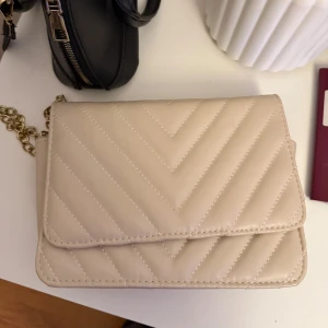 Beige quiltad axelväska med kedja - Säljer en stilren beige axelväska med quiltat mönster och guldfärgad kedja. Väskan har flera fack inuti och stängs med magnetknapp. Perfekt för att bära det viktigaste och passar till många outfits.