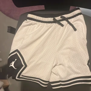 Vita Jordan Dri-Fit shorts med svart kant - Snygga vita Jordan shorts med svart kant och klassisk Jumpman-logga på benen. Tillverkade i Dri-Fit material som andas och har snörning i midjan. Perfekta för träning eller sportiga outfits.