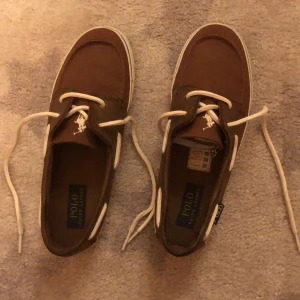 Bruna seglarskor från Polo Ralph Lauren - Bruna seglarskor från Polo Ralph Lauren med vit snörning och vit sula. Skorna har klassisk design med broderad logga på ovansidan. Perfekta för en avslappnad stil.Priset är  hugget i sten . 