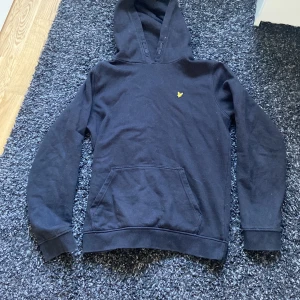 Svart hoodie från Lyle & Scott Junior - Säljer en svart hoodie från Lyle & Scott Junior med klassisk gul logga på bröstet. Tröjan har huva och en stor magficka framtill. Perfekt för en avslappnad och stilren look.
