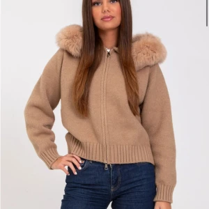 Beige stickad jacka med fuskpäls - Säljer en beige stickad jacka med dragkedja och stor huva med fluffig fuskpäls. Jackan har ribbade muddar och en avslappnad passform, perfekt för kyliga dagar.