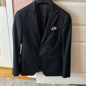 Kostym från Riley - Säljer en svart kostym från Riley i slim fit-modell. Kavajen har två knappar, klassiska slag och en bröstficka. Tillhörande byxor med enkel bakficka. Perfekt för formella tillfällen. Kostymen är bara använd en gång och skicket är 10/10. Byxorna är st 44 och kavajen st 46.Nypris på kostymen på brothers-2699, mitt pris 1400. Hör av er vid mer frågor.