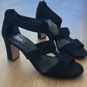 Svarta klackade sandaler från So All - Säljer ett par svarta sandaler med klack från So All. Skorna har öppen tå, breda remmar och dragkedja bak. Klacken är stabil och i trälook, vilket ger en snygg kontrast till det svarta materialet.