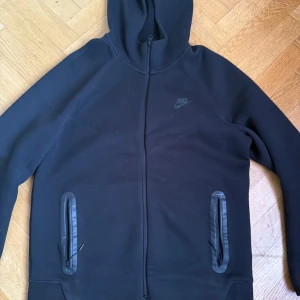 Svart hoodiejacka från Nike - Svart hoodiejacka från Nike med dragkedja framtill och två fickor med dragkedja. Jackan har huva och diskret Nike-logga på bröstet. Perfekt för en sportig och avslappnad stil.