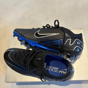 Nike Mercurial fotbollsskor svart/blå - Säljer ett par Nike Mercurial fotbollsskor i svart med blå och vita detaljer. Skorna har snörning, rund tå och platt sula. Perfekta för dig som vill ha lätta och snabba skor på planen. Snygg design med Nike-logga på sidan. Påse ingår!