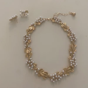 Guldfärgat halsband och örhängen med pärlor - Säljer ett set med halsband och örhängen i guldfärgat metall med dekorativa blad och vita pärlor. Halsbandet har fina detaljer med bladformade länkar och pärlor längs hela kedjan. Örhängena matchar med samma blad och pärldesign. Halsbandets modell är inte långt utan det är ett ganska kort halsband som är super snyggt! Priset går att diskuteras💞