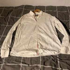 Vit skjorta från Massimo Dutti - Säljer en klassisk vit skjorta från Massimo Dutti med snygga detaljer i form av röd och blå rand längs knappslån. Skjortan har lång ärm och är tillverkad i ett mjukt bomullstyg. Perfekt för dig som gillar stilrena plagg med en twist.