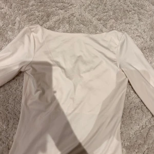 Vit långärmad topp med djup ringning - Säljer en stilren vit långärmad topp med djup rundad ringning framtill. Toppen är figurnära och tillverkad i ett mjukt bomullsliknande material. Perfekt att matcha med jeans eller kjol.