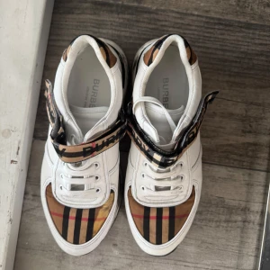 Burberry vita sneakers med klassiskt rutmönster - Säljer ett par vita sneakers från Burberry med det ikoniska beige, svarta och röda rutmönstret. Skorna har snörning och en bred rem med Burberry-logga över vristen. Perfekta för dig som vill sticka ut med lyxig streetstyle.