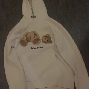 Vit hoodie från Palm Angels med nallemotiv - Säljer en vit hoodie från Palm Angels med ett unikt nallemotiv på framsidan. Hoodien har en klassisk design med dragsko i huvan och ribbade muddar vid ärmslut och nederkant. Perfekt för en avslappnad stil.