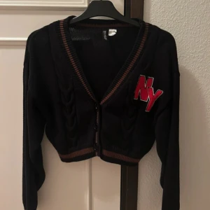 Kofta - Säljer en svart kofta från H&M Divided med röd 'NY' applikation på bröstet i storlek S. Koftan har knappar framtill och ribbade detaljer i brunt och rött vid ärmslut och nederkant. Perfekt för en casual look!