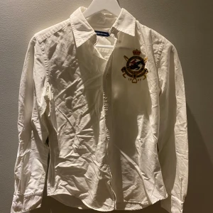 Vit skjorta från Ralph Lauren Sport - Snygg vit skjorta från Ralph Lauren Sport med broderad emblem på bröstet. Skjortan har långa ärmar och klassisk krage. Perfekt för en stilren look. Passar S