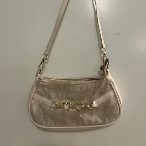 Beige axelväska med hjärtformade detaljer - Säljer en söt beige axelväska med guldfärgade hjärtformade detaljer på framsidan. Väskan har en dragkedja upptill och en justerbar axelrem. Den är dock lite gammal och inte i perfekt skick men fungerar som nytt ändå