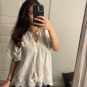 Vit blus med spetsdetaljer - Charmig vit blus med vackra spetsdetaljer och korta ärmar. Blusen har en lös passform och knytning vid halsen. Perfekt för en somrig look.