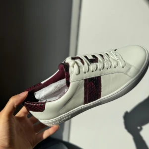 Vita sneakers från Michael Kors - Snygga vita sneakers från Michael Kors med vinröda ormskinnsdetaljer på sidan och hälen. Skorna har klassisk snörning och en stilren design som passar perfekt till vardags. HELT NYA