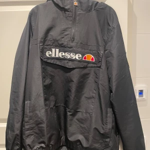 Svart vindjacka från Ellesse - Säljer en svart vindjacka från Ellesse med dragkedja och stor ficka framtill. Jackan har en luva och elastiska muddar vid ärmslut och nederkant. Perfekt för blåsiga dagar.