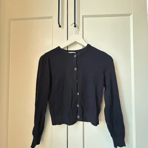 Mörk blå kofta med knappar - Säljer en stilren svart kofta med knappar framtill. Den har långa ärmar och en klassisk rund halsringning. Perfekt för lager-på-lager under kyligare dagar.