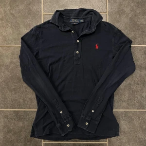 Svart långärmad pikétröja från Ralph Lauren - Säljer en stilren svart långärmad pikétröja från Ralph Lauren med den klassiska röda loggan broderad på bröstet. Tröjan har knappar vid kragen och är perfekt för en avslappnad men ändå elegant look.