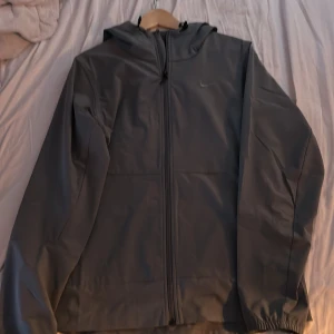 Grå Nike Windbreaker - Säljer en stilren grå vindjacka från Nike med dragkedja och huva. Perfekt för blåsiga dagar. Jackan har en diskret Nike-logga på bröstet och är tillverkad i ett lätt syntetmaterial.