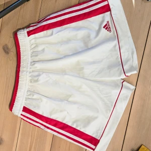 Vita shorts från Adidas med röda detaljer - Säljer ett par vita Adidas shorts med röda ränder och logga. De har en elastisk midja och är perfekta för sportiga aktiviteter. Klassisk design med tre ränder längs sidorna.