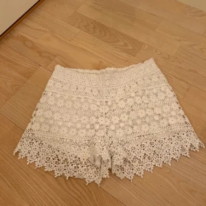 Vita spets shorts - Säljer ett par vita shorts med vacker spetsdesign. De har en högmidjad passform och är perfekta för sommarens varma dagar. Shortsen har en fin franskant som ger en extra touch av elegans.