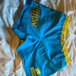 Blå badshorts från NOCCO - Snygga blå badshorts från NOCCO med gul elastisk midja och snörning. Perfekta för sommarens badäventyr. Har en bakficka med NOCCO-logga.