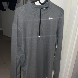 Grå långärmad tröja från Nike - Säljer en grå långärmad tröja från Nike med dragkedja vid halsen. Perfekt för träning eller vardagsbruk. Den har en enkel och stilren design med Nike-loggan på bröstet. Som ny då jag använt 5 gånger efter köp storleken är L men sitter mer åt M hållet