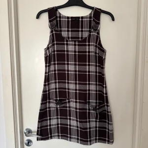 Brun och vit rutig klänning från Eve - Snygg brun och vit rutig klänning från Eve i storlek XS. Klänningen har breda axelband och dekorativa knappar framtill. 