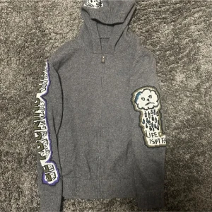 Grå hoodie med tryck - Säljer en grå hoodie med dragkedja och unikt tryck på ärmarna och bröstet. Designen inkluderar text och illustrationer i olika färger. Perfekt för en avslappnad stil.