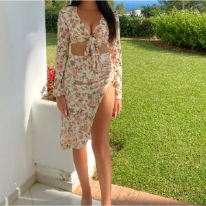 Blommig klänning med knyt och slits - Säljer en vacker blommig klänning med knyt framtill och en djärv slits. Klänningen har långärmade ärmar och en omlott-design som ger en elegant look. Perfekt för sommarens festligheter!