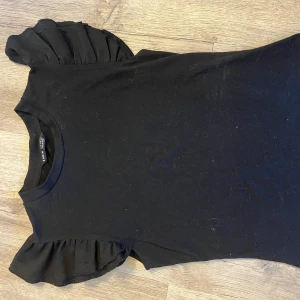 Svart topp med volangärmar från Shein - Säljer en svart topp från Shein med korta volangärmar. Perfekt för en stilren look med en twist. Toppen är i ett mjukt material och har en rund halsringning.