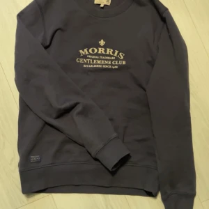 Sweatshirt från Morris - Säljer en stilren mörkblå sweatshirt från Morris med tryck på bröstet. Tröjan har långa ärmar och rund halsringning. Perfekt för en avslappnad och trendig look.