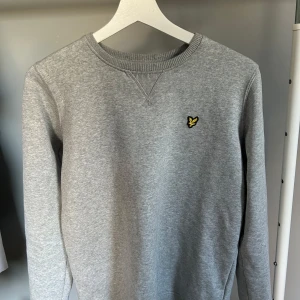 Grå sweatshirt från Lyle & Scott - Säljer en stilren grå sweatshirt från Lyle & Scott med deras ikoniska gula logotyp på bröstet. Tröjan har en rund halsringning och ribbade muddar vid ärmslut och nederkant. Perfekt för en avslappnad look.