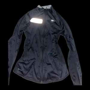 Svart träningsjacka från Nike - Snygg svart träningsjacka från Nike med dragkedja och långärmad design. Perfekt för löpning eller gymmet. Jackan har en reflekterande detalj på bröstet och är tillverkad i ett lätt och andningsbart material.