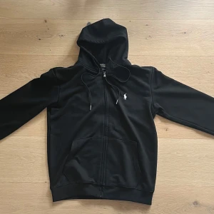 Svart hoodie från Polo Ralph Lauren - Säljer en stilren svart hoodie från Polo Ralph Lauren med dragkedja och broderad logga på bröstet. Hoodien har långa ärmar och en praktisk huva med snörning. Perfekt för en avslappnad look.