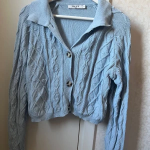 Ljusblå stickad kofta från NA-KD - Säljer en ljusblå stickad kofta från NA-KD med kabelstickat mönster och stora knappar framtill. Koftan har en klassisk krage och långa ärmar❤️