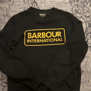 Svart tröja från Barbour International - Säljer en svart tröja från Barbour International med ett gult tryck på framsidan. Tröjan har långa ärmar och en rund halsringning. Perfekt för en stilren och avslappnad look.