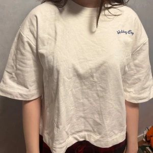 Vit t-shirt med tryck 'Holiday Explore' - Säljer en vit t-shirt med blått tryck 'Holiday Explore' på både fram- och baksidan. T-shirten har en avslappnad passform och korta ärmar, perfekt för en ledig stil. Den har aldrig använts och har prislappen kvar. Den är lite för stor på mig som är S men borde passa perfekt på M💕