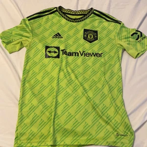 Grön Manchester united tröja från Adidas - Säljer grön Manchester united fotbollströja från Adidas med svarta detaljer och tryck. Tröjan har korta ärmar och ett mönster över hela tyget. Den har ett klubbmärke på bröstet och spelarens namn och nummer på ryggen.
