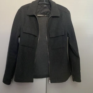 Svart jacka från H&M - Snygg overshirt från H&M med dragkedja framtill och två bröstfickor. Jackan har en klassisk krage och långa ärmar, perfekt för en stilren look. Passar bra till både vardag och mer uppklädda tillfällen. Använt bara ett fåtal gånger storlek S men passar mera som M