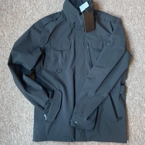 Snoot Livorno fieldjacka - Snygg svart jacka från Snoot med flera praktiska fickor och dragkedjor. Jackan har en stilren design med hög krage och knappar på axlarna. Perfekt för en modern look. Kostar runt 5 tusen i butik. Helt ny.