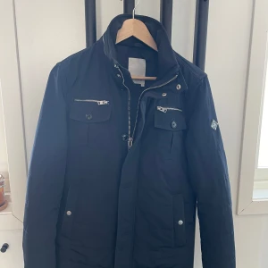 Jacka från J.Lindeberg - Snygg svart jacka från J.Lindeberg med dragkedja och knappar. Jackan har flera praktiska fickor med dragkedjor och en stilren design. Perfekt för kyligare dagar.