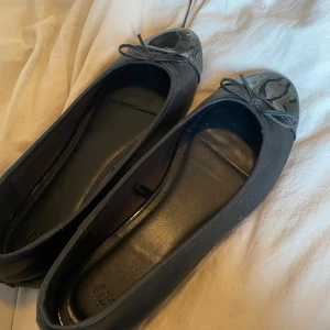 Svarta ballerinaskor med rosett - Snygga svarta ballerinaskor med en elegant rosett på tån. Skorna har en glansig finish på tån som ger en stilren look. Perfekta för en chic och bekväm stil.