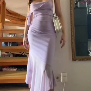 Ljuslila klänning med volangdetalj - Säljer en elegant ljuslila klänning med smala axelband och volangdetalj framtill. Klänningen har en figurnära passform och en vacker volangkant vid fållen. Perfekt för sommarens festligheter!