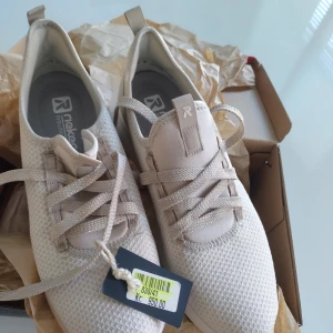 Beige sneakers från Rieker - Snygga och bekväma beige sneakers från Rieker med snörning och en mjuk sula för extra komfort. Skorna har en stilren design med en vit och beige yttersula som ger bra grepp. Perfekta för en avslappnad stil.