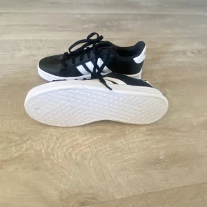 Svarta och vita sneakers från Adidas - Snygga svarta sneakers med vita detaljer från Adidas. Skorna har klassisk snörning och en vit sula. Perfekta för en stilren look.