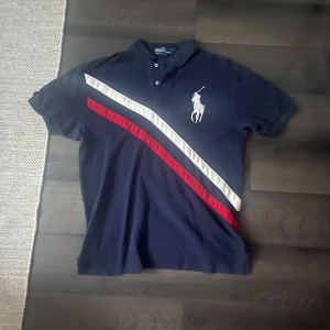 Ralph Lauren piké  - Snygg marinblå pikétröja från Polo Ralph Lauren med vita och röda diagonala ränder över bröstet. Klassisk krage, knappar framtill och stor broderad logga. Perfekt för dig som gillar sportig och stilren look. Axelbredd 50cm längd 68cm. Jag skickar ej bild när jag bär plaggen och jag vet ej vilken längd och vikt de passar då jag inte prövar kläderna jag säljer, så för storlek hänvisar jag till måtten och passform att man googlar på modellnamn etc 😃